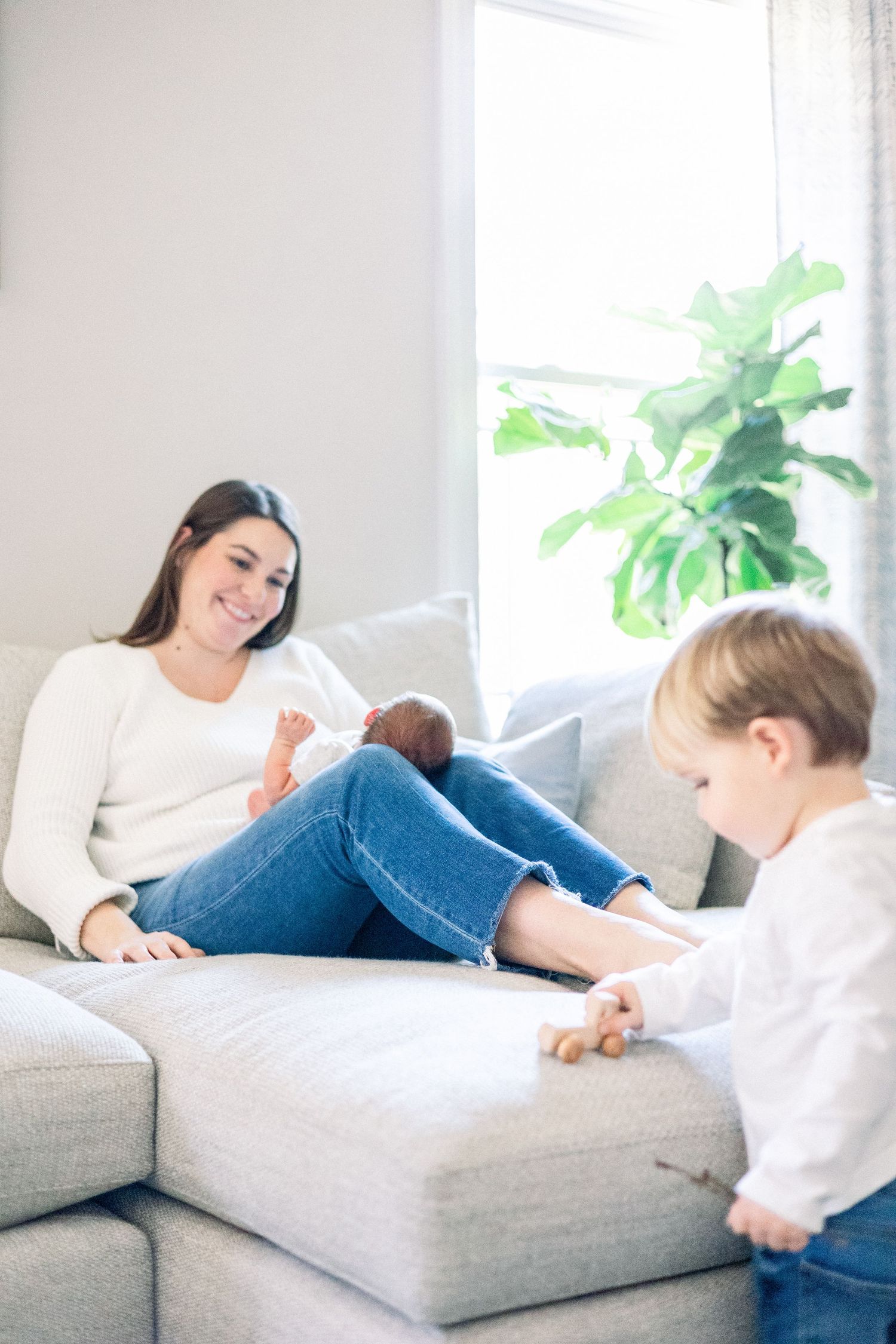 A mother and child share a tender moment together on a light gray couch next to a window with a green plant. Evans newborn photographer.
