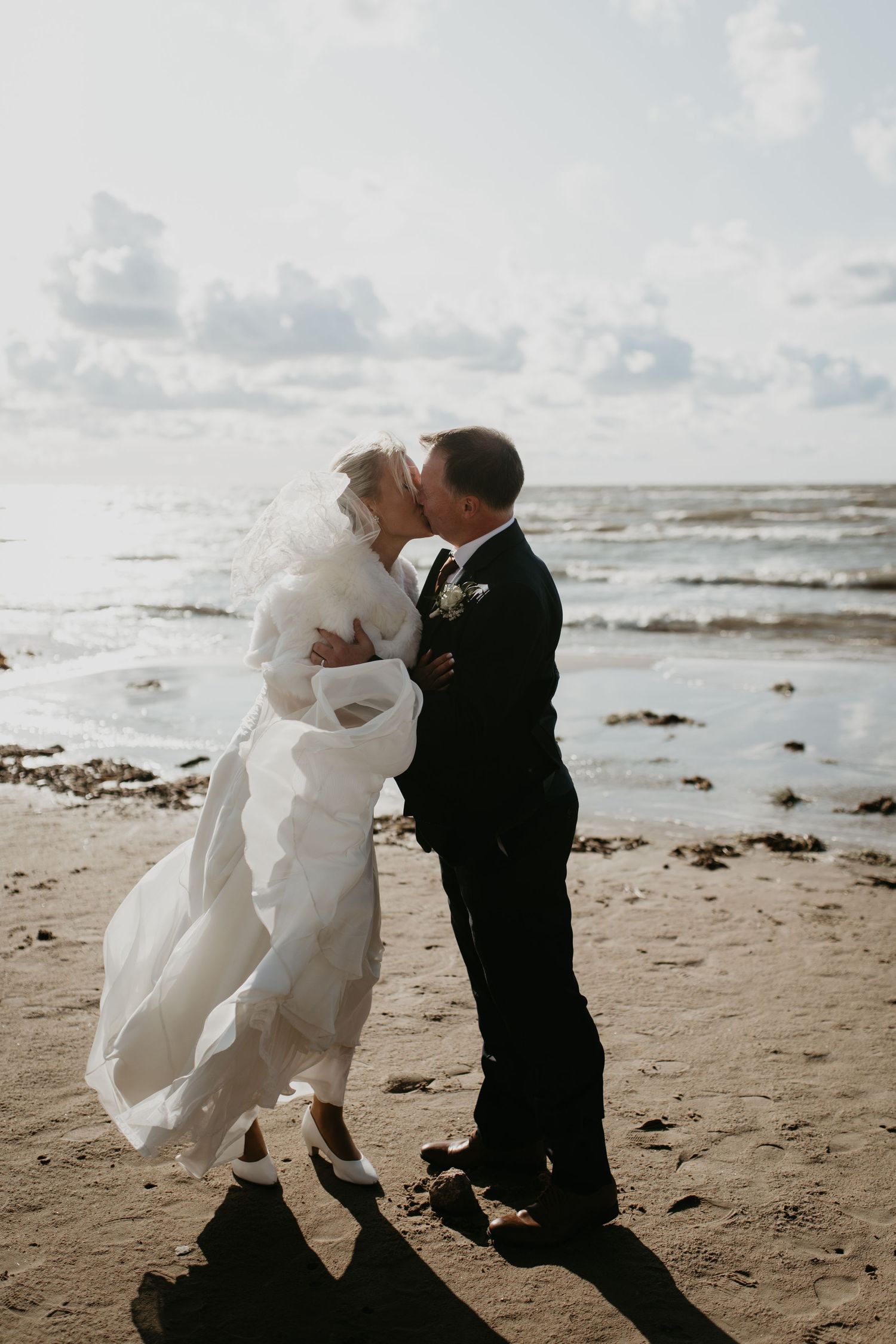 Two people share a romantic moment on a windy beach at sunset with waves and cloudy sky in the background.