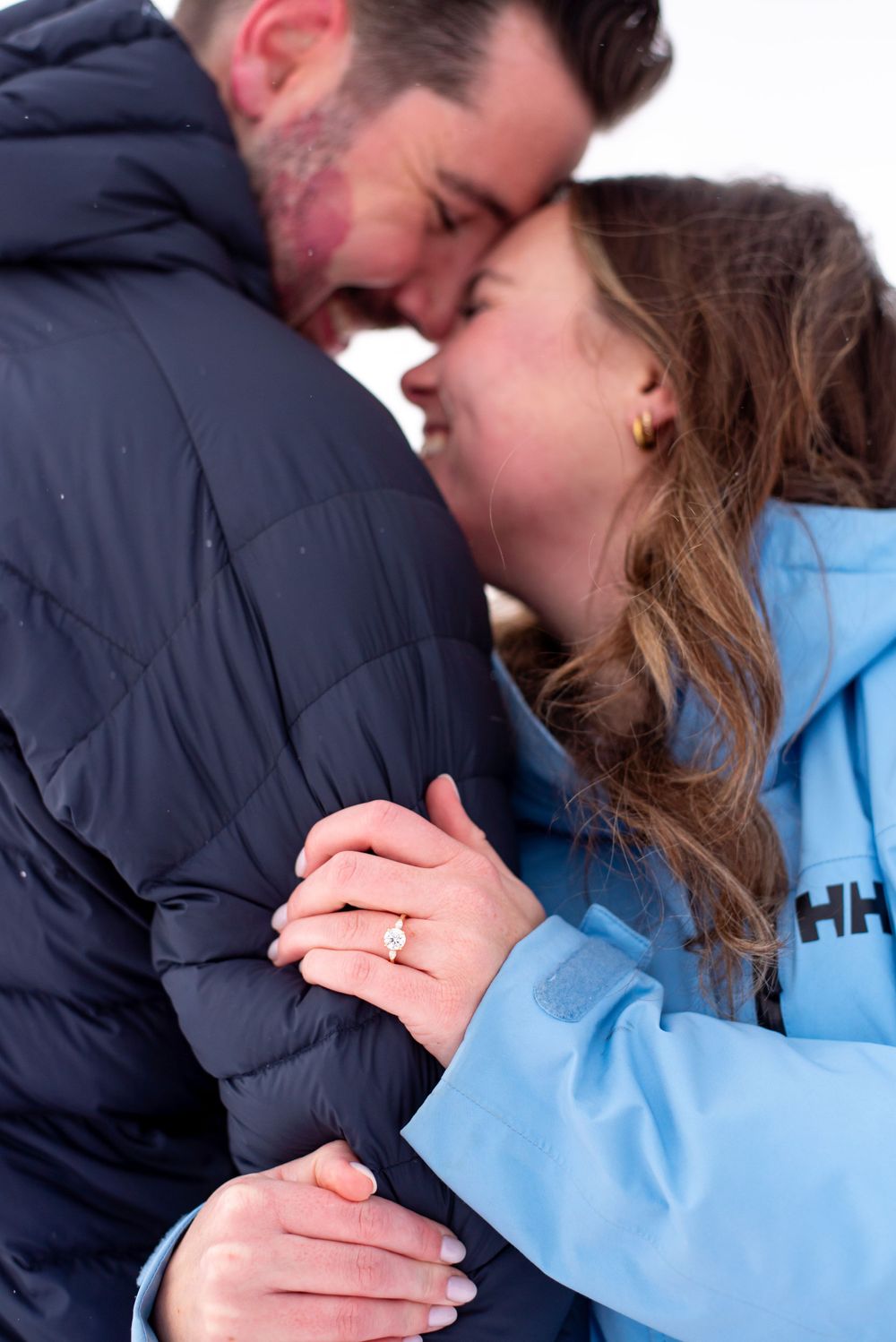 An engaged couple in winter coats sharing an intimate moment with a ring visible.