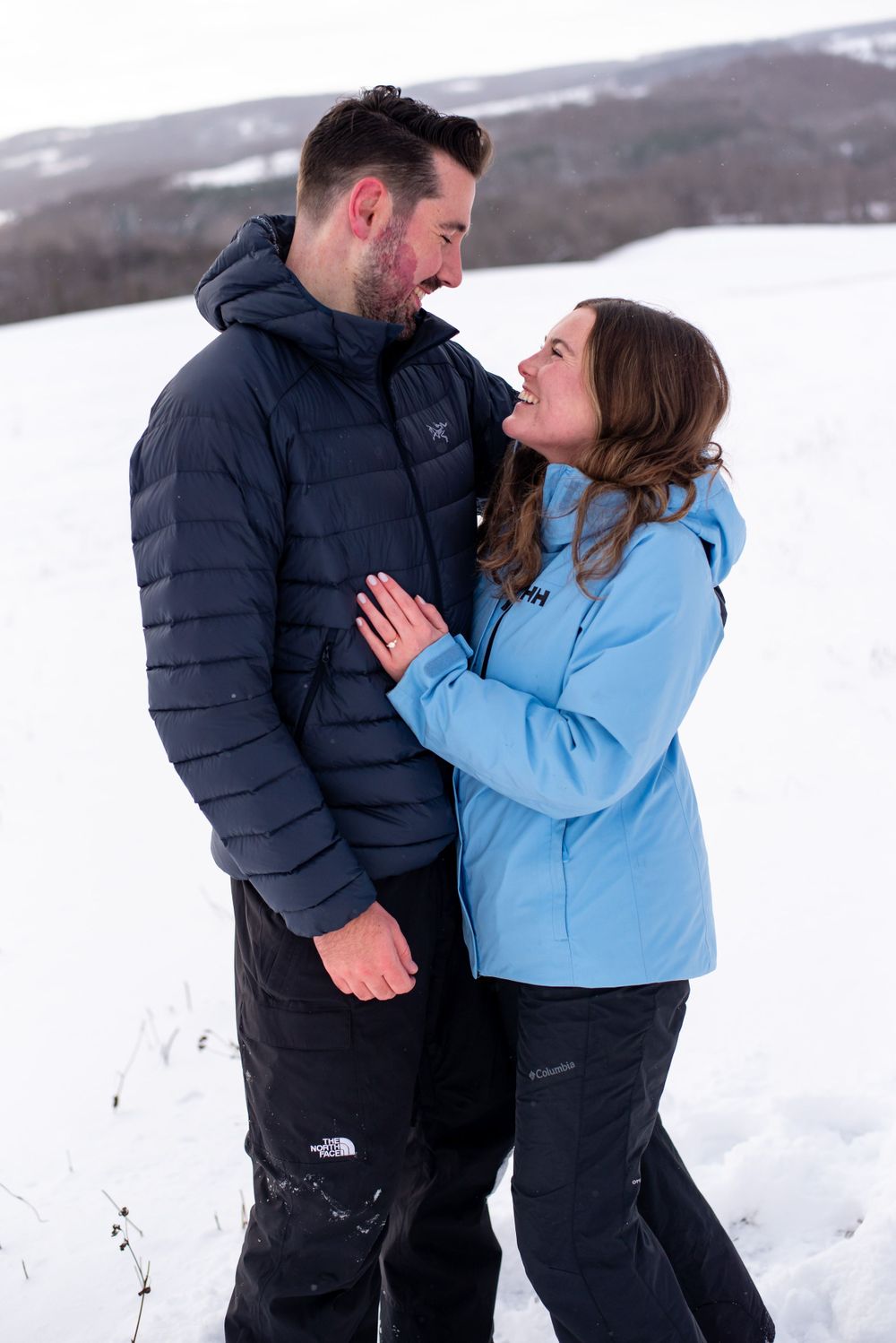 A couple laughing together in winter coats against a snow-covered landscape.