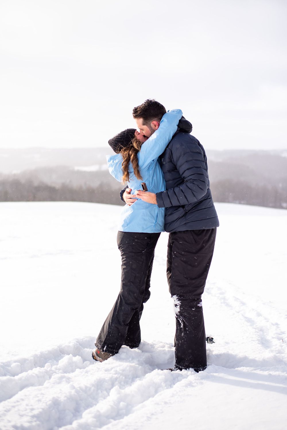 Two people in winter coats share an intimate embrace while standing in deep snow on a misty winter day.
