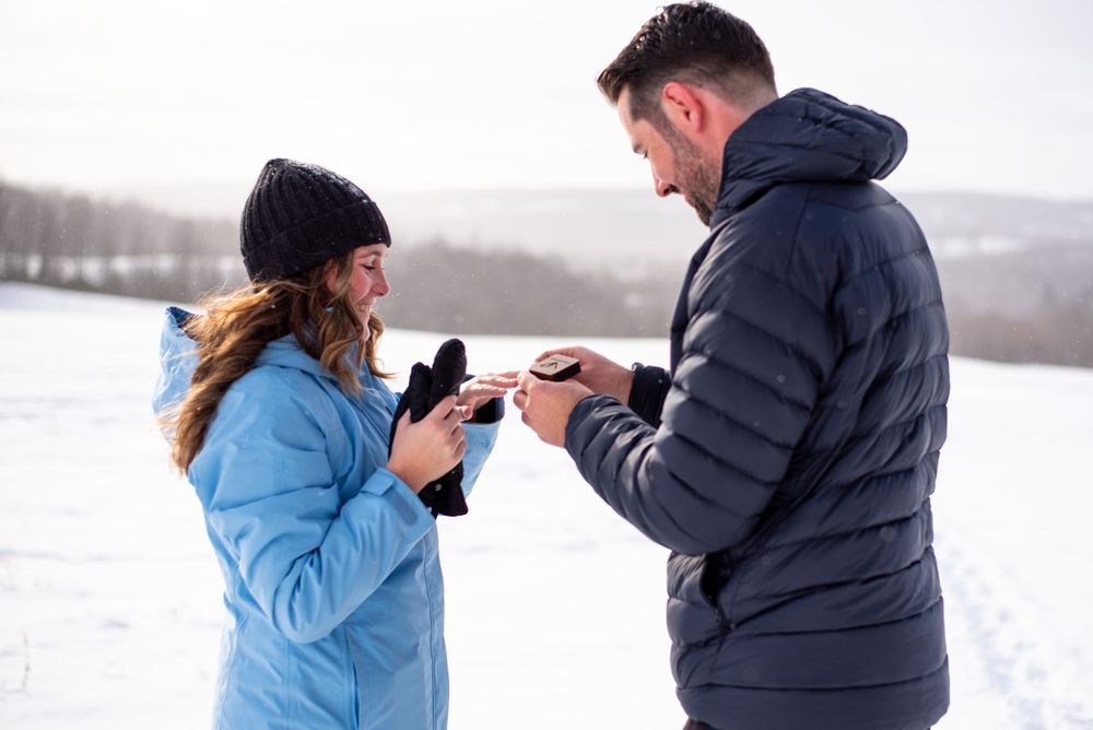 A man in a winter jacket opens an engagement ring box for a woman in a winter jacket, in a snowy field on a cloudy day.