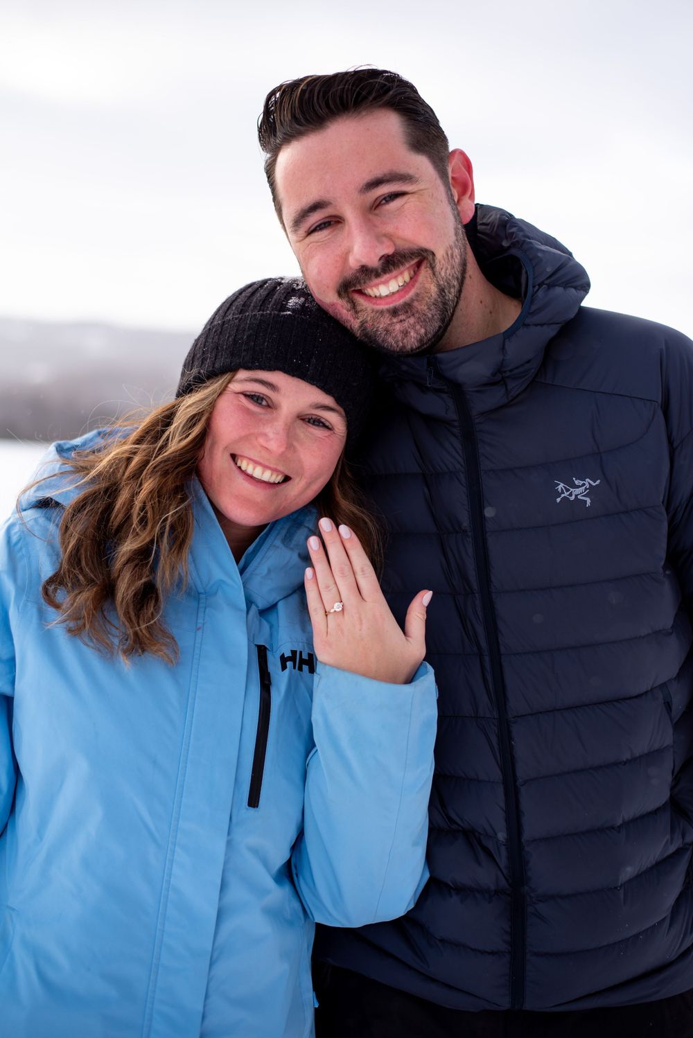 A happy couple in winter jackets showing off an engagement ring in the snow.