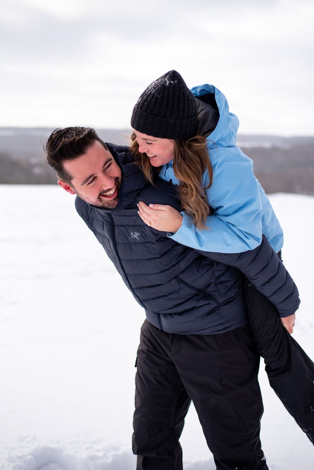 A couple shares a playful piggyback ride moment in a snowy winter landscape.