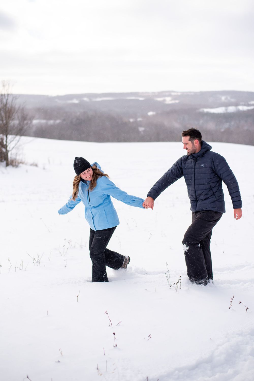 A couple holds hands while walking through deep snow with rolling hills in the background.