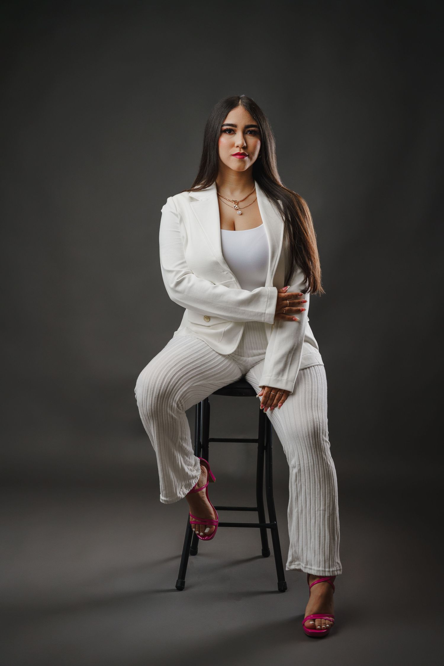 Professional portrait of someone in an all-white business suit and pink heels posing on a stool against a dark background.