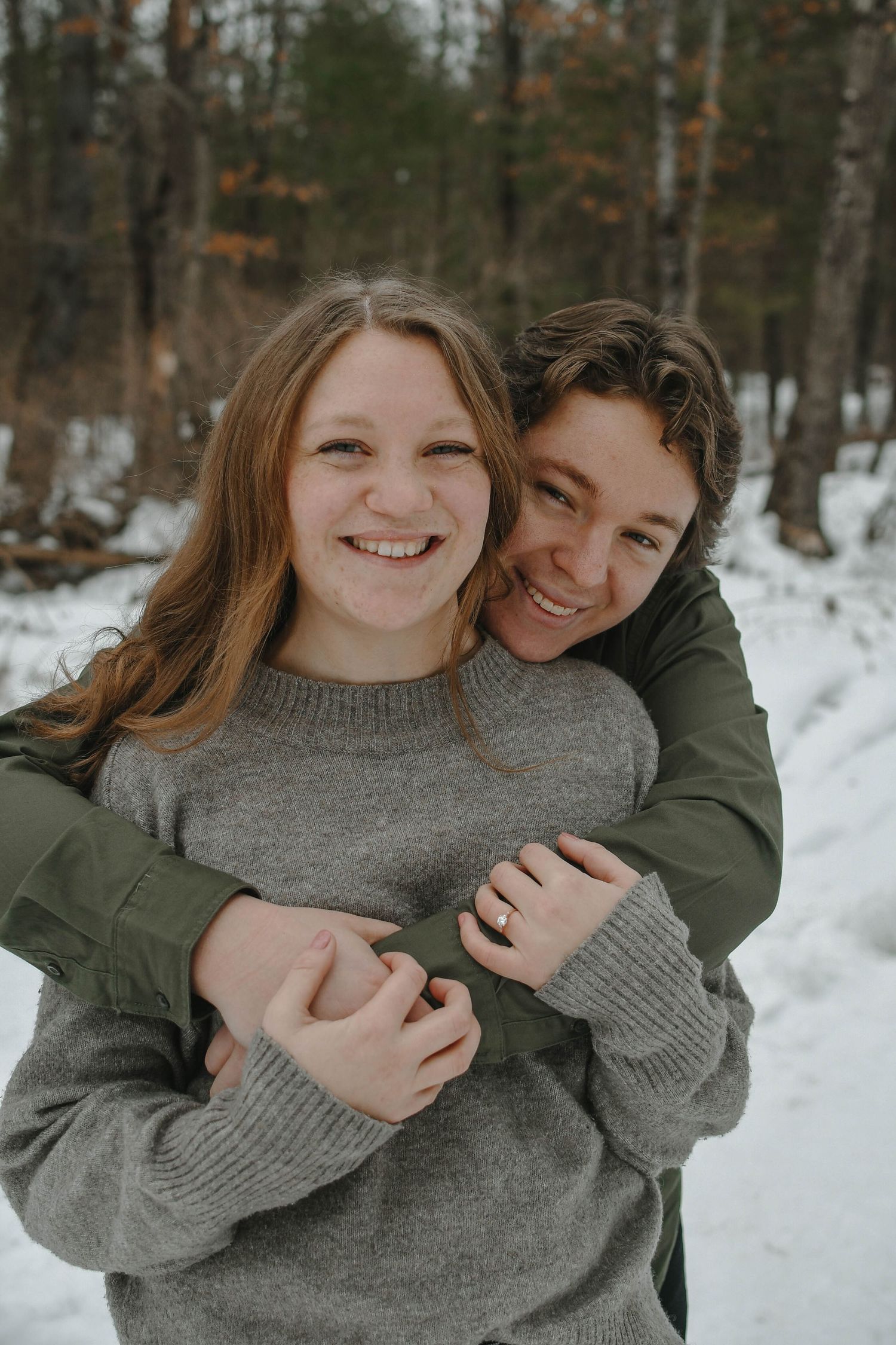 A couple embraces in a snowy winter forest setting wearing casual sweaters and sharing a joyful moment together.