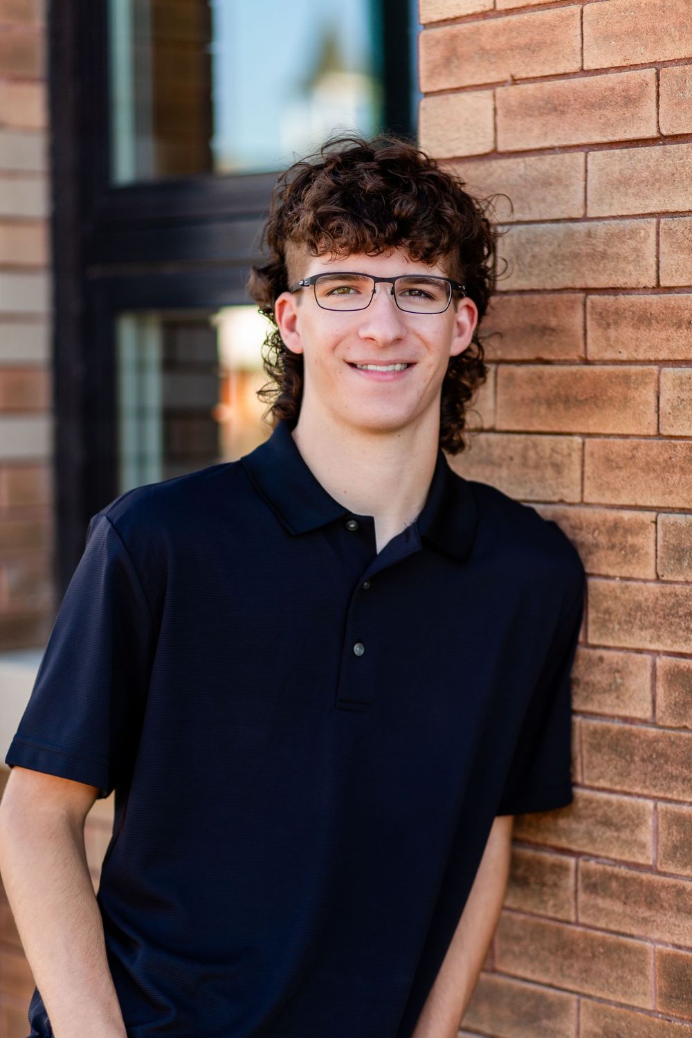 A young person with curly hair and glasses wearing a navy polo shirt stands against a brick wall smiling.