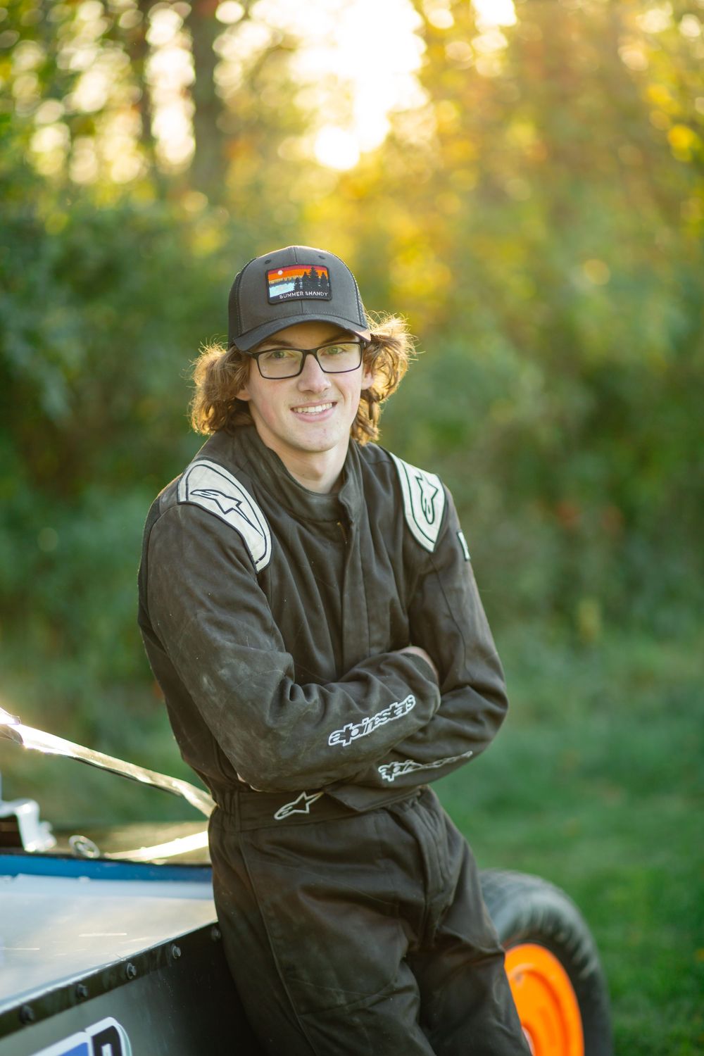A figure wearing glasses and a baseball cap sits outdoors at sunset with a warm golden glow in the background.