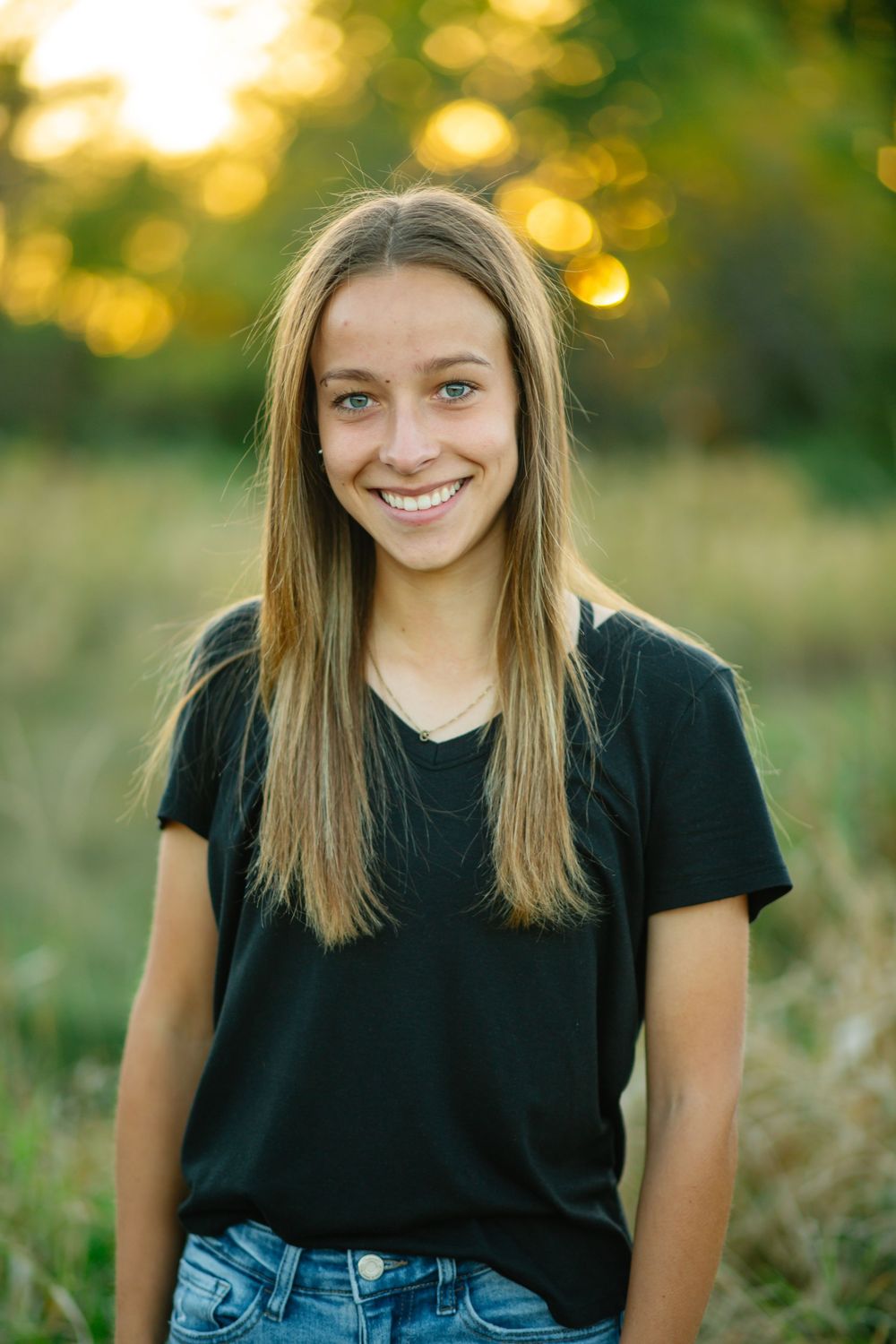 Young person in black t-shirt and jeans sitting outdoors with golden bokeh sunlight in background during portrait session.