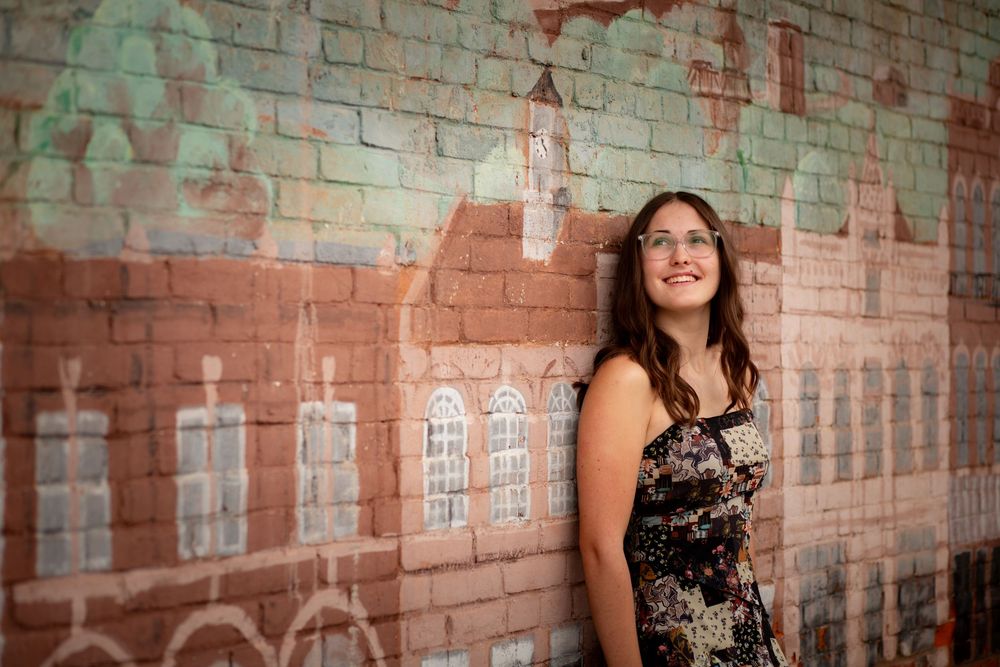 A woman in a floral dress stands against a weathered brick wall with faded painted designs in natural lighting.