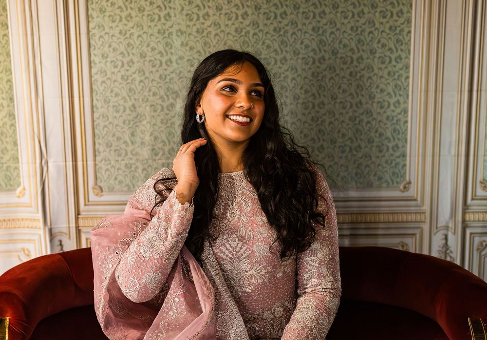 Woman wearing cozy pink sweater sitting on grey chair and smiling warmly while posing for portrait.