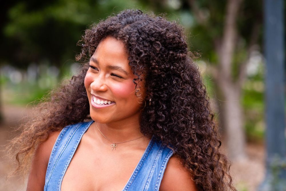 Woman with curly hair and denim top smiles joyfully while posing outdoors in natural lighting.