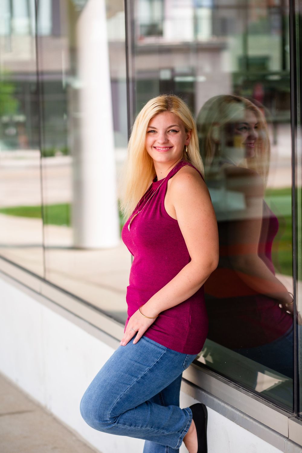 Someone wearing a burgundy sleeveless top and blue jeans poses against a window in a modern building.