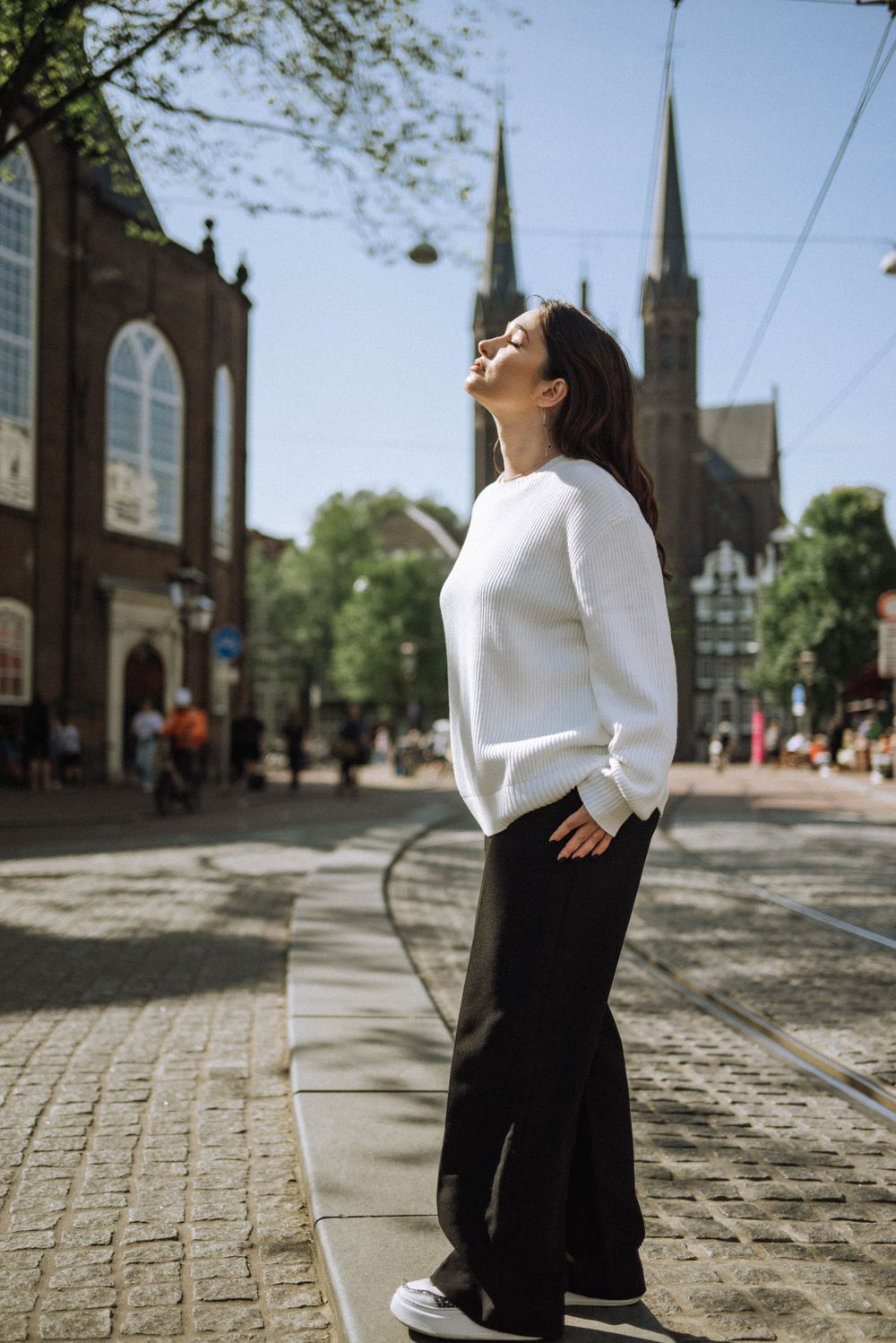 Person in white sweater and black pants posing on cobblestone street with church spires in background.
