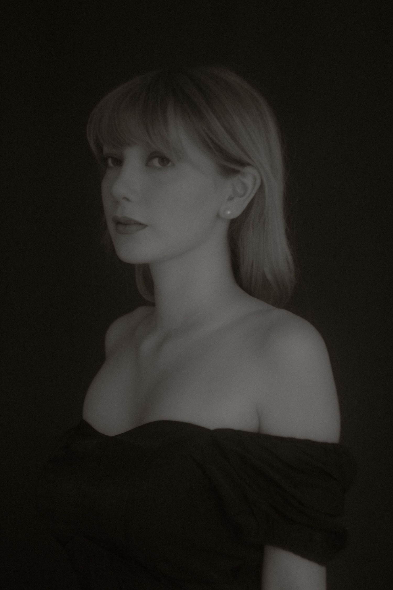 Elegant black and white portrait of blonde woman in off-shoulder top gazing upward on dark background.
