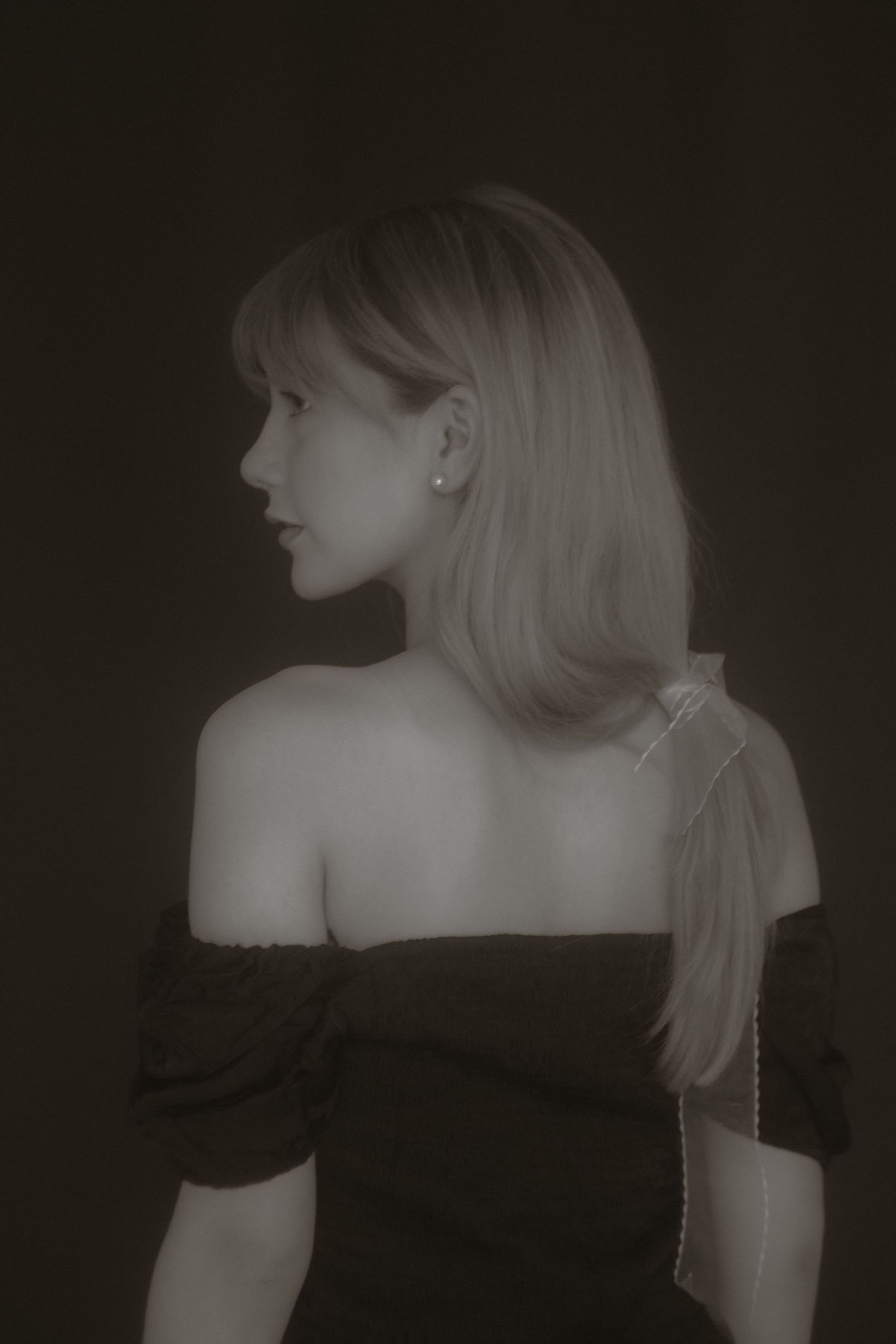 Black and white portrait of blonde woman in off-shoulder top viewed from behind on dark background.
