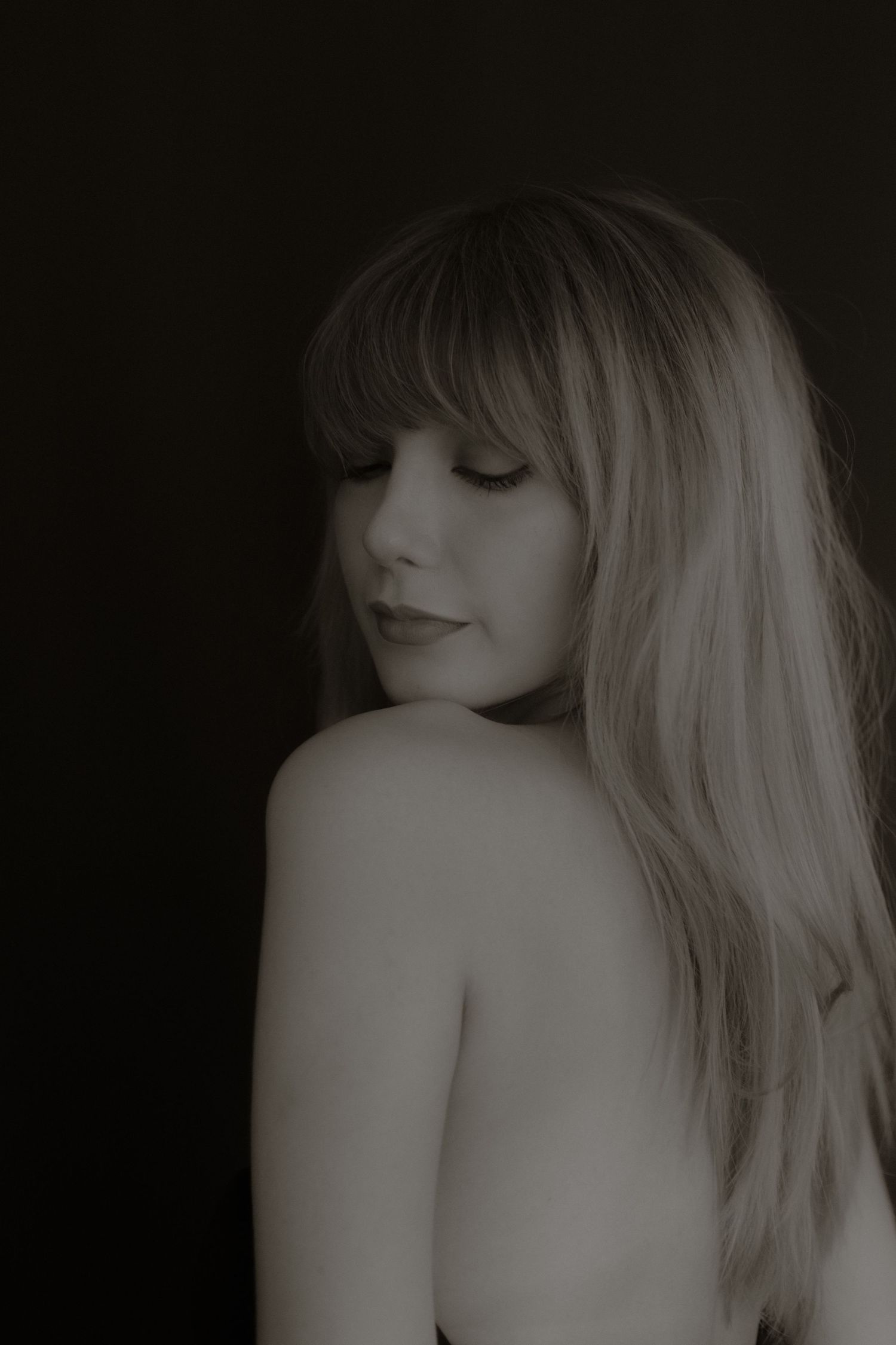 Soft black and white portrait of blonde woman with bangs and bare shoulder on dark background.