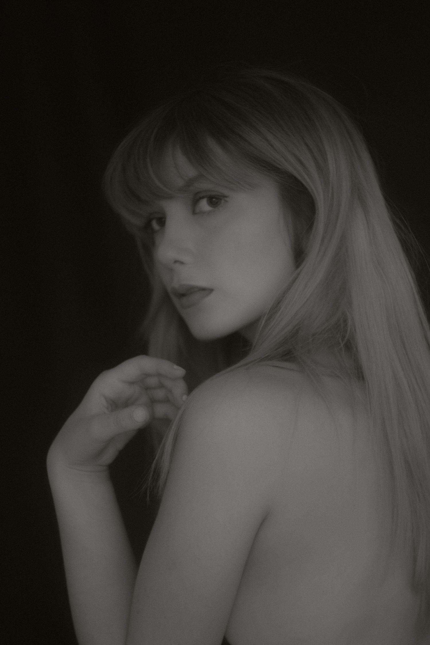 Moody monochrome portrait of a blonde woman with bangs, bare shoulders, dark background.