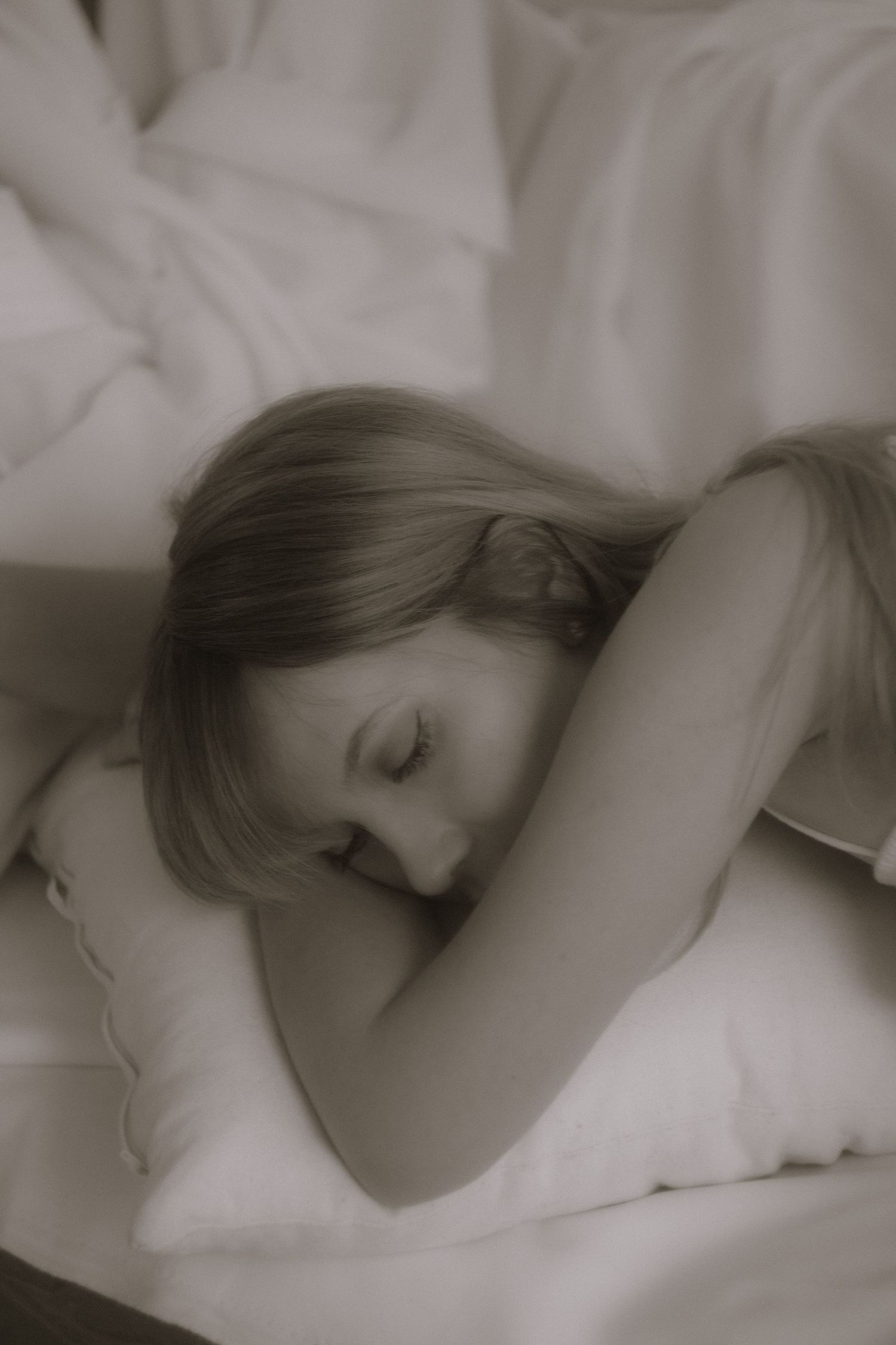 Soft sepia overhead shot of a blonde woman resting on white pillows in bed.