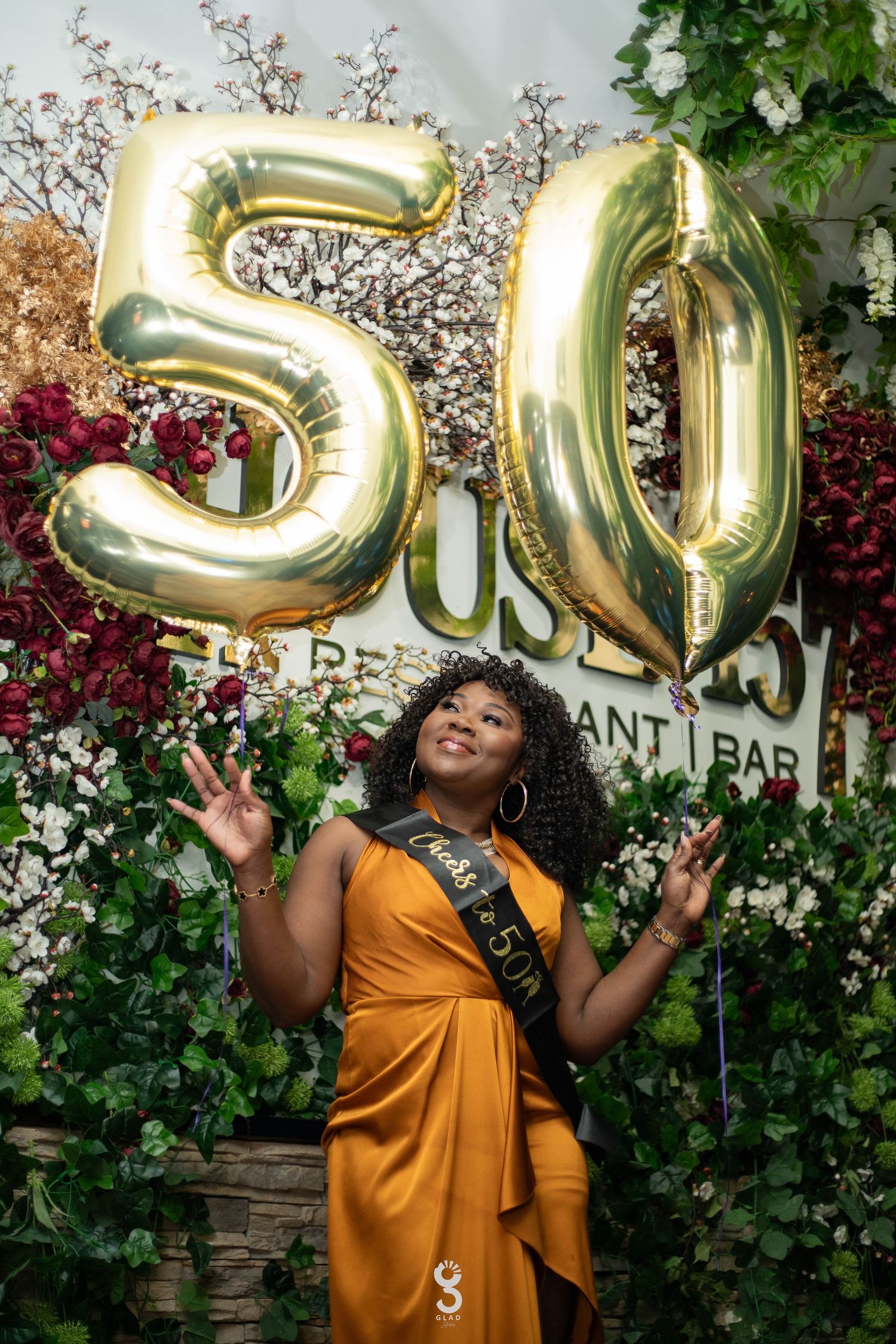 A festive 50th birthday celebration setup with large gold number balloons and lush greenery decorations in a garden setting.