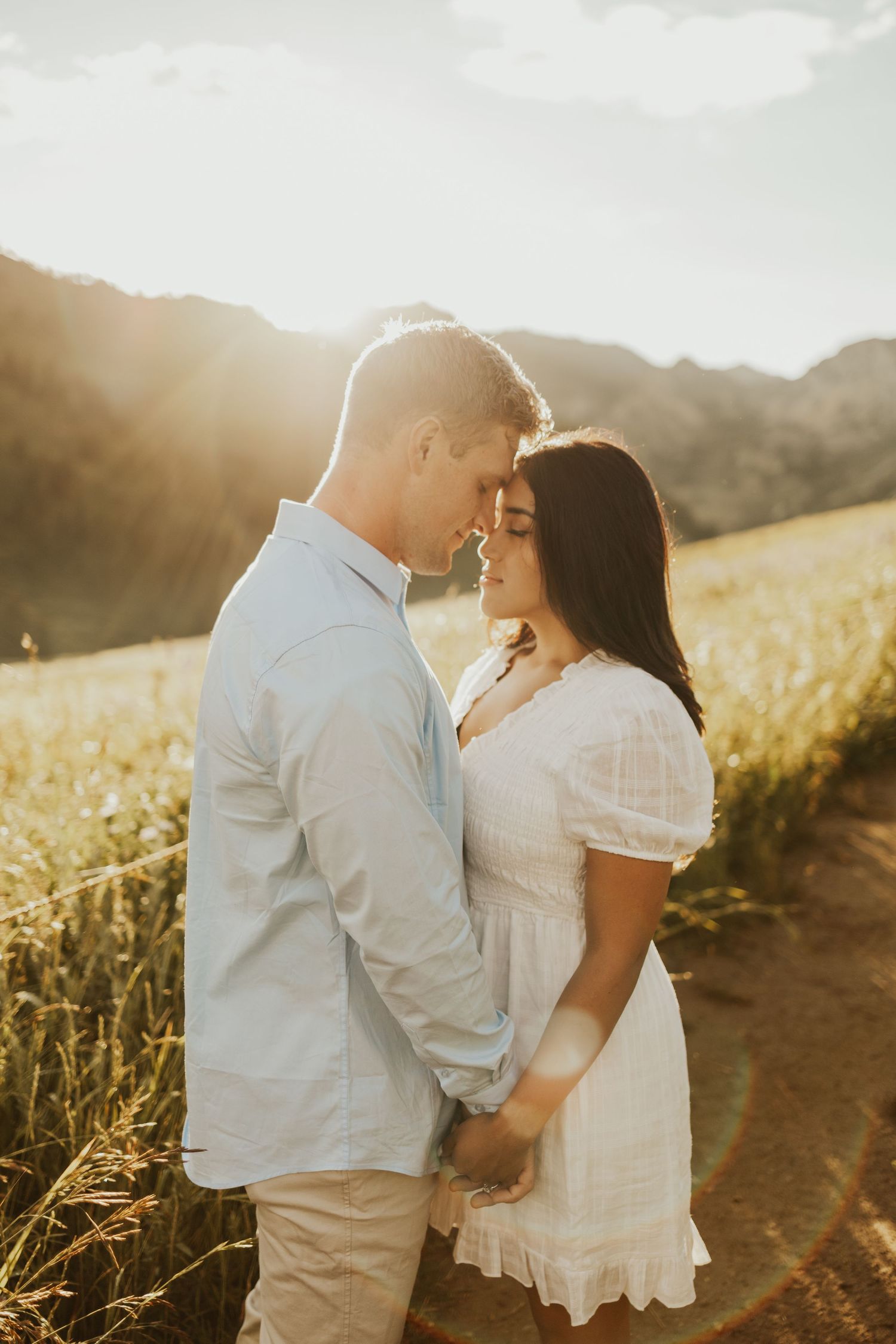 Couple holding hands and touching foreheads in golden sunlit field with mountain backdrop.