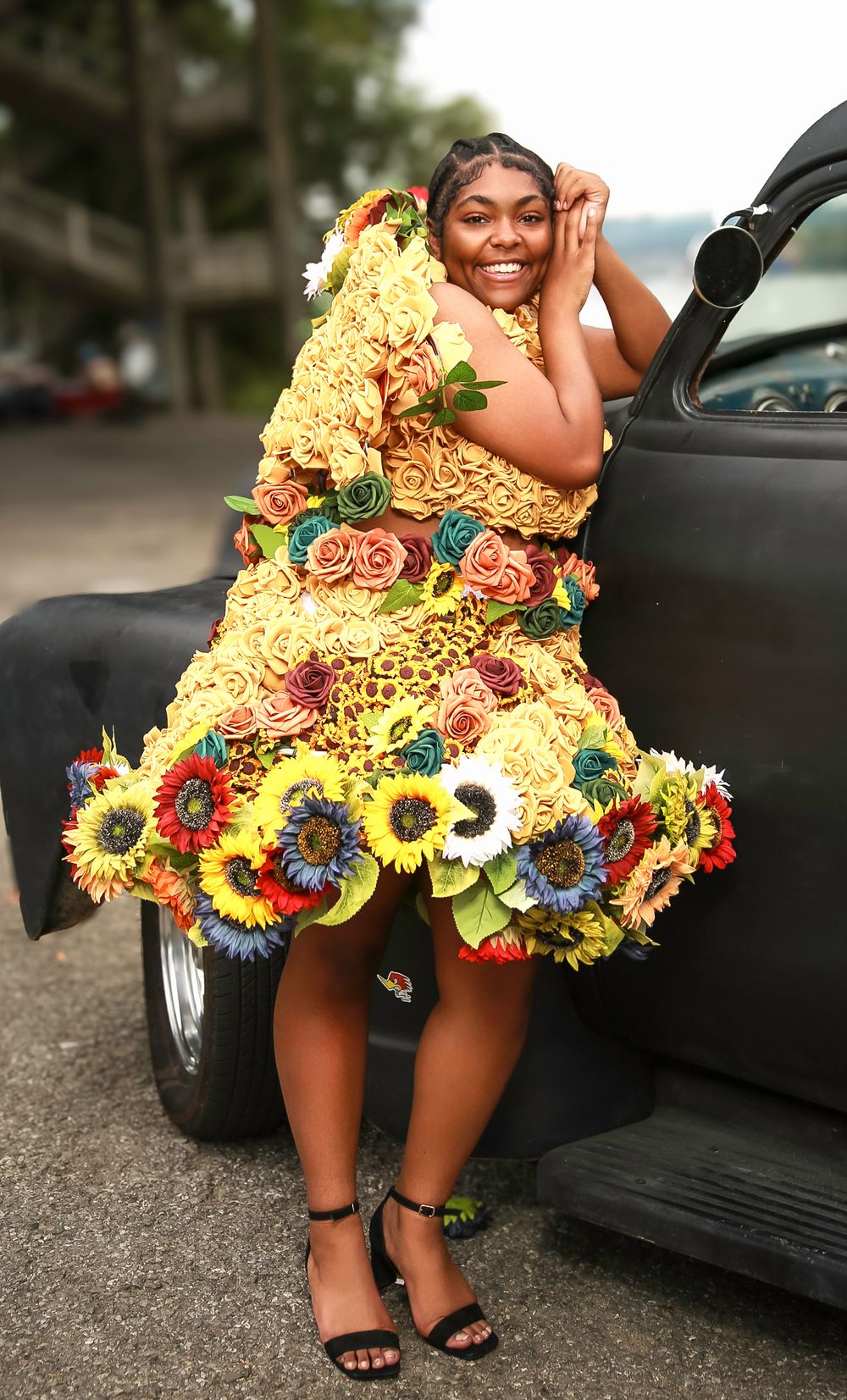 A person wearing a vibrant yellow dress adorned with colorful sunflowers and roses poses next to a black car.