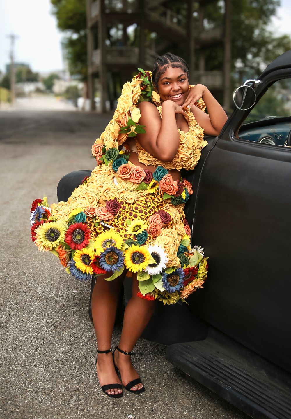 A vibrant yellow floral dress adorned with sunflowers and colorful blooms stands out against a car and street backdrop.