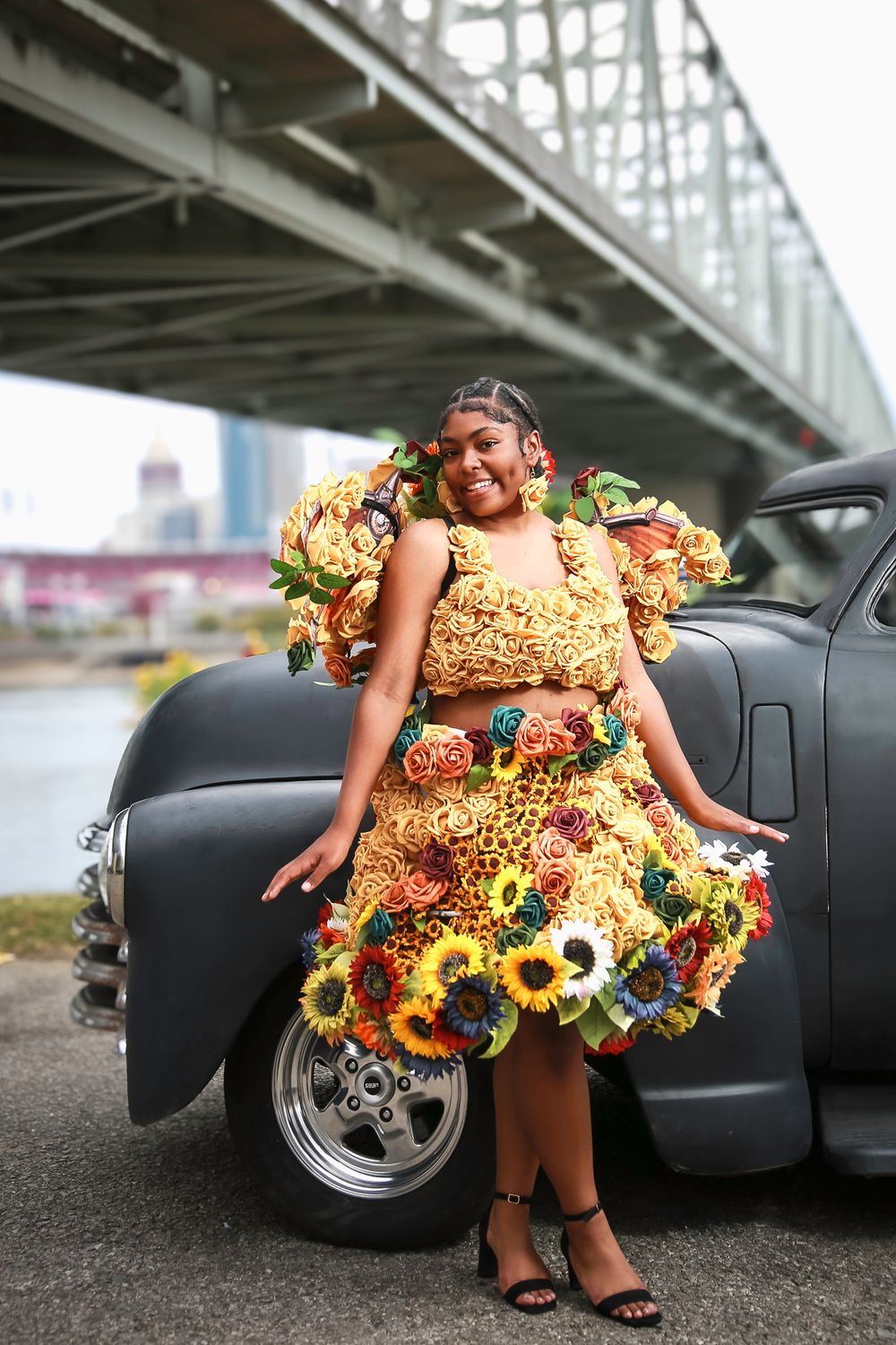 Colorful fashion design featuring a dress adorned with vibrant sunflowers posed near a vintage car under a bridge.