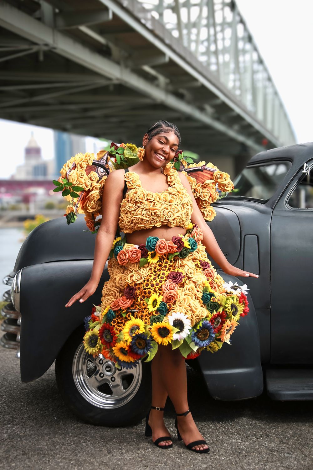 Joyful fashion photograph showing a sunflower-decorated dress against a classic black car backdrop under a bridge.