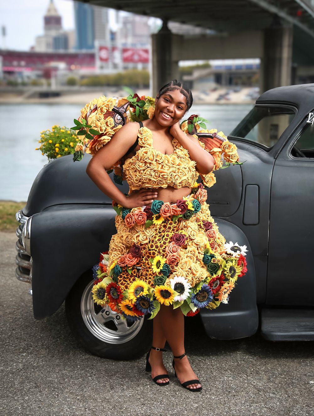 A model poses in a vibrant dress made of flowers and floral patterns next to a vintage black car outdoors.
