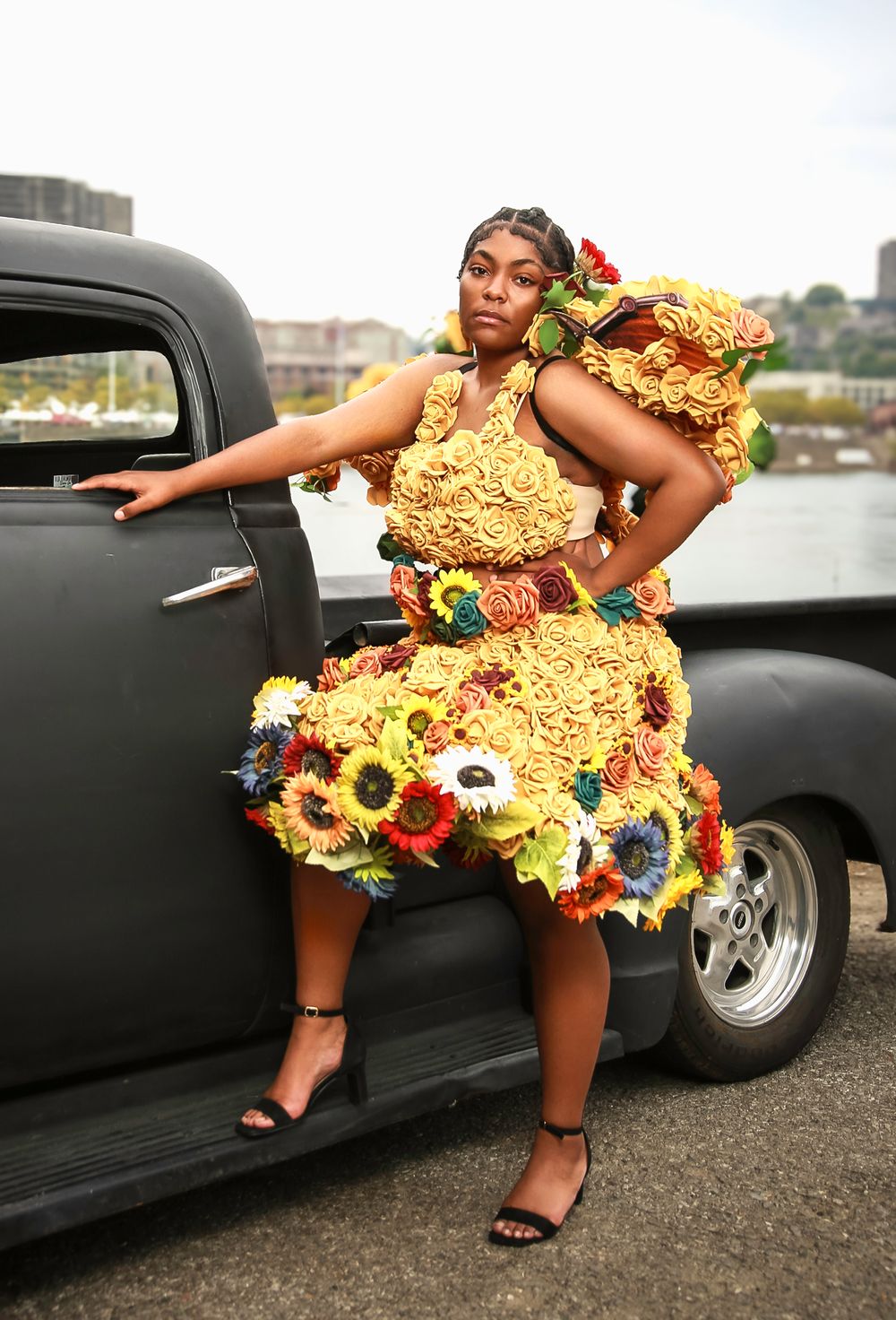Creative fashion portrait featuring a dress made entirely of ramen noodles adorned with colorful flowers next to a vintage car.
