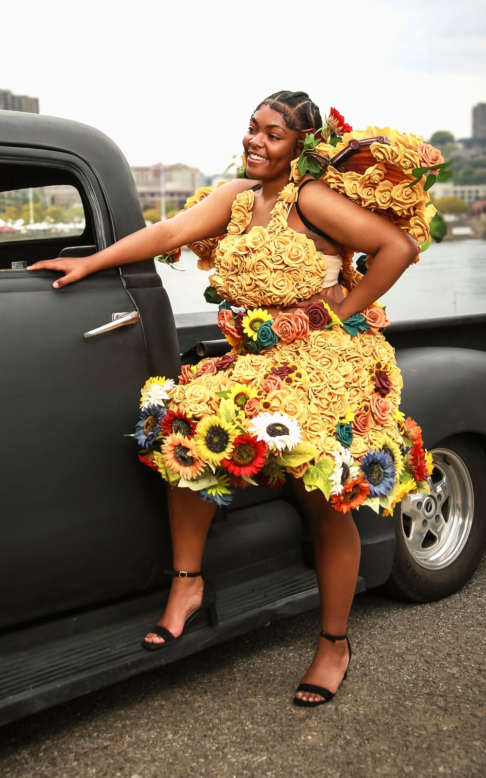 A person in a vibrant yellow dress adorned with sunflowers poses against a classic black car at an outdoor location.