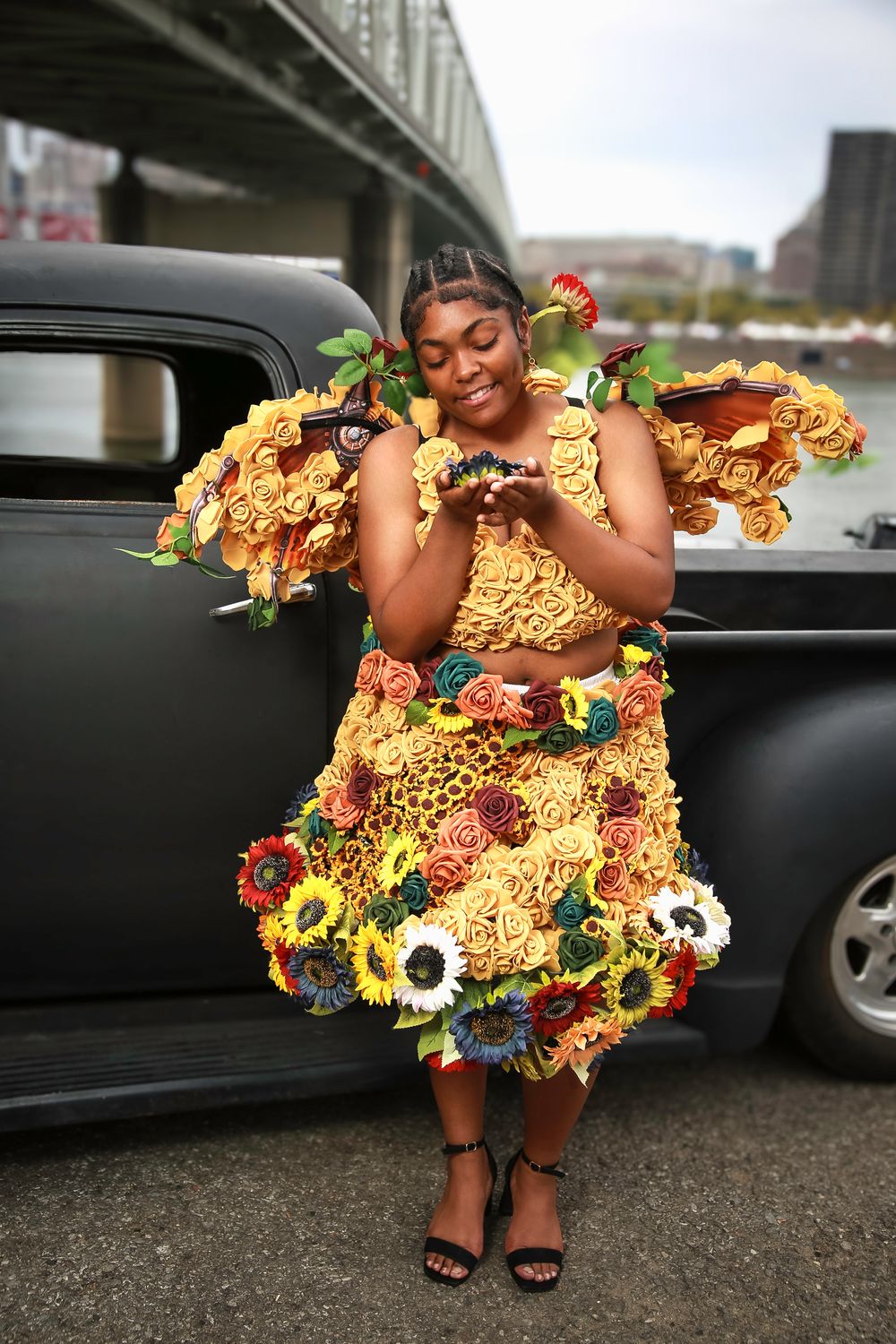 A creative dress made entirely of ramen noodles and colorful decorations worn in front of a vintage black car.