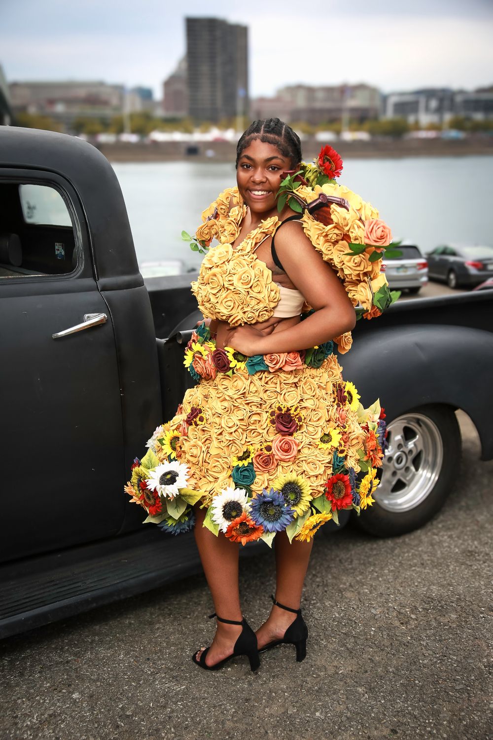 Someone poses by a vintage black car wearing a unique yellow dress decorated with colorful flowers at a waterfront location.