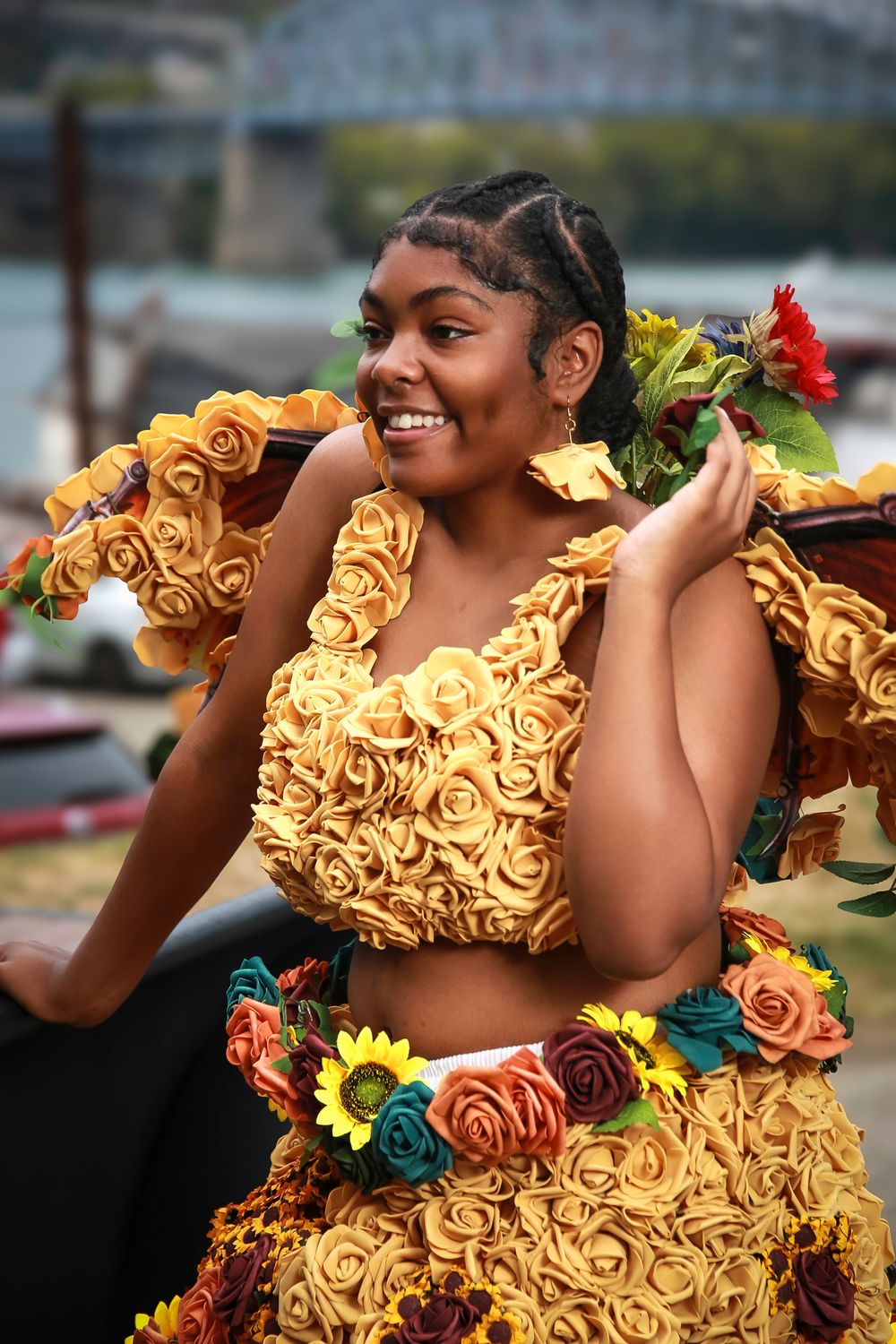 A person wearing an ornate yellow floral dress with colorful bouquet accents poses outdoors with a bright smile.