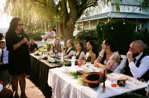 A long outdoor dining table set with white linens and glassware under trees during a garden party gathering.