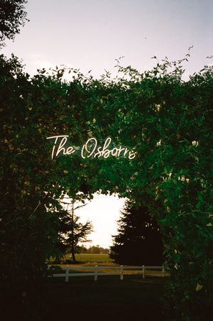 A rustic outdoor sign reading 'The Osborns' illuminated at sunset with lush green foliage framing the scene.