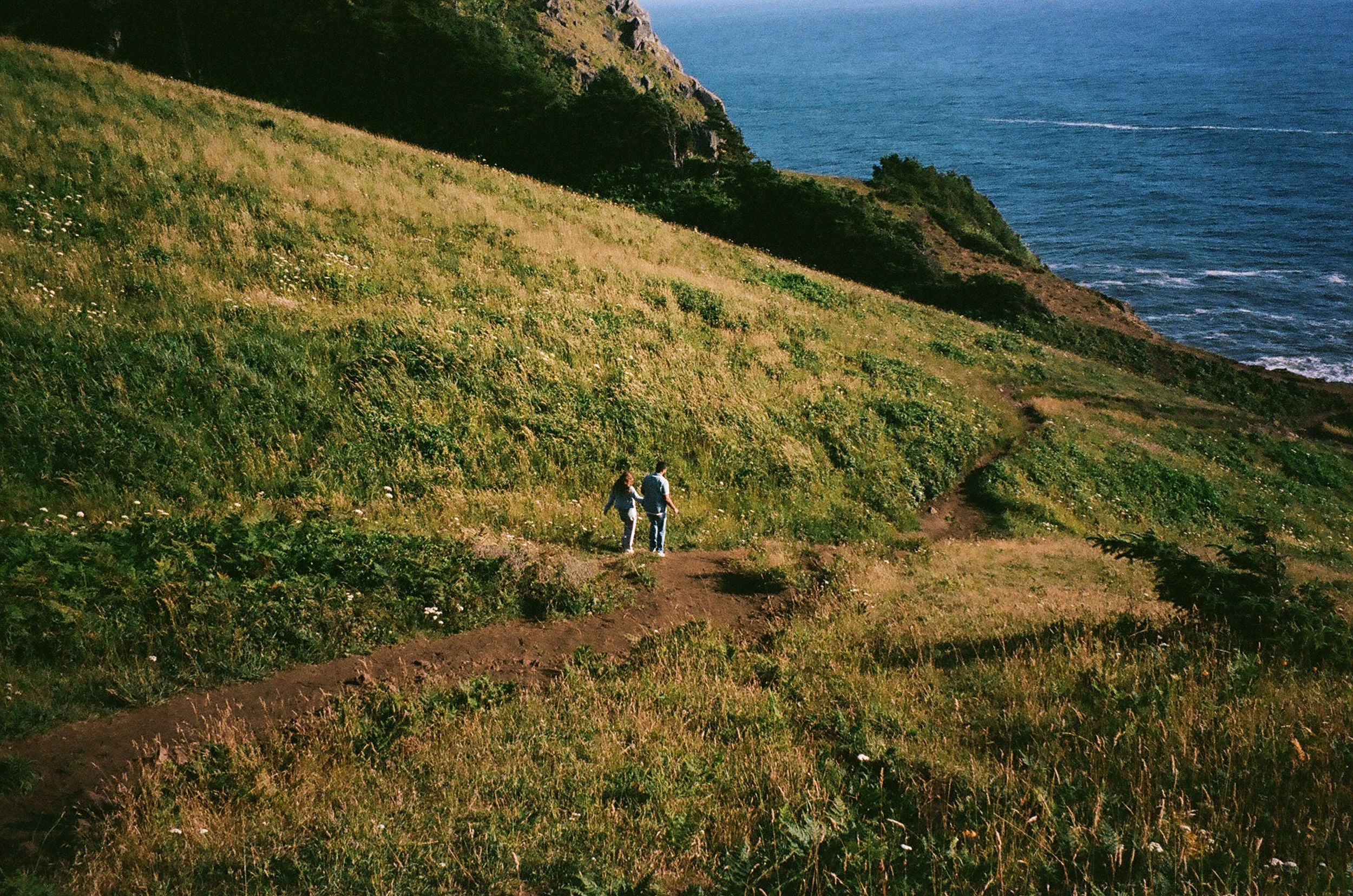 A hiker walks along a coastal trail on a grassy cliff overlooking the blue ocean on a sunny day.