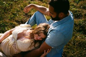 A couple rests together on grass in warm sunlight, with one person wearing a lace dress and the other in denim.