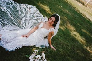A bride in a flowing white lace wedding dress poses gracefully on a grassy lawn in natural sunlight.