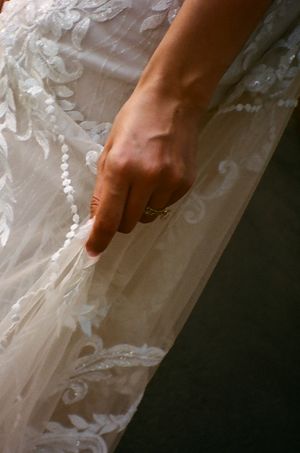 A hand wearing an engagement ring gently touches intricate lace details on a white wedding dress.