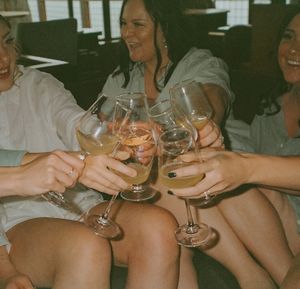 Friends clink wine glasses together in celebration while sitting and enjoying drinks at a gathering.