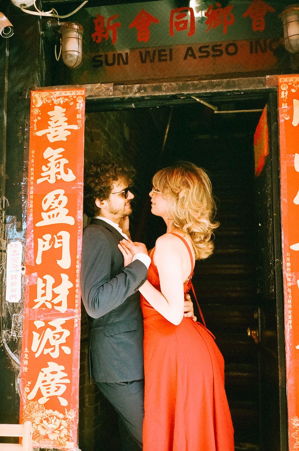 Couple in elegant attire embracing in a doorway adorned with red Chinese decorative panels and Sun Wei Asso sign.