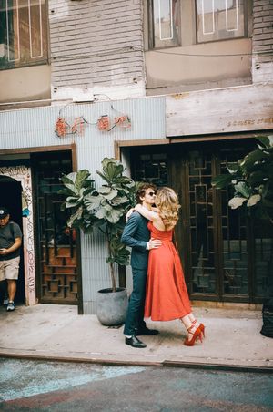 Couple hugging on a Chinatown street, woman in red midi dress and heels, man in a blue suit.