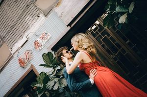 Couple embracing and kissing outdoors, woman in elegant red dress and man in navy suit surrounded by tropical foliage.