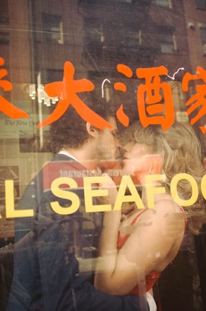 Couple kissing in front of a Chinese seafood restaurant window with red Chinese characters reflected in the glass.