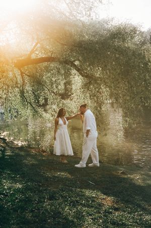 Couple in white outfits sharing a romantic moment under a willow tree by a sunlit river.