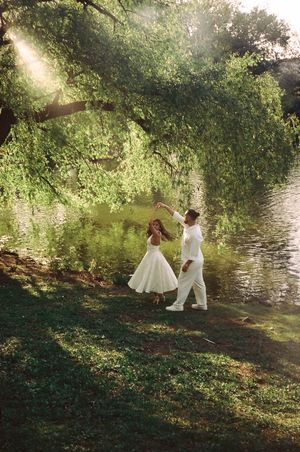 Couple in white outfits dancing by a sunlit green lake under a large tree.