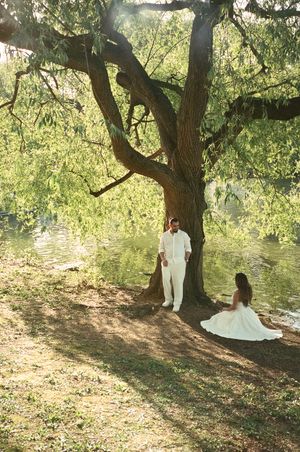 Couple in white wedding attire share a romantic moment under a large tree by a sunlit pond.